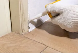 Gloved hand sealing floor gap with caulking gun.
